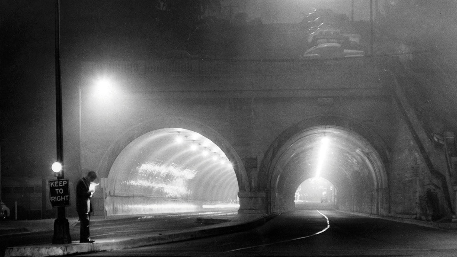 A black and white photo of a man stood underneath a streetlight, with two tunnels in the background