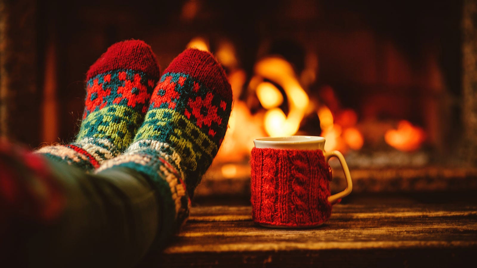 A pair of feet in warm Christmassy socks, beside a mug wrapped in cosy, both in front of a roaring fire.