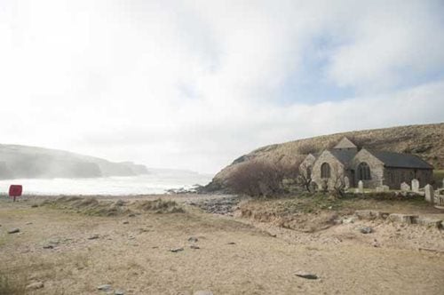 A beach on a cloudy day, with a small church to the left and side and mist over the bay