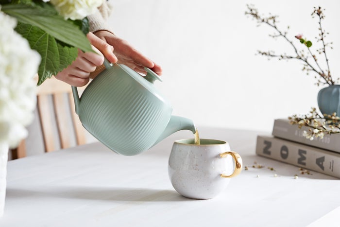 tea being poured into a white cup with a gold handle, from a green teacup, with a stack of books topped by dried flowers in the background