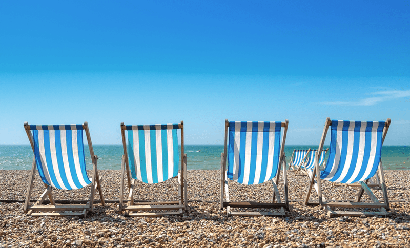 Four blue and white striped deckchairs on a pebble beach facing the sea and a blue sky 