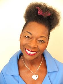 Photograph of Floella Benjamin smiling, wearing a blue shirt and a red bow in her hair