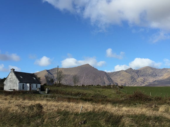 small white cottage in a rugged field with long grasses and mountains in the background, with a blue sky above