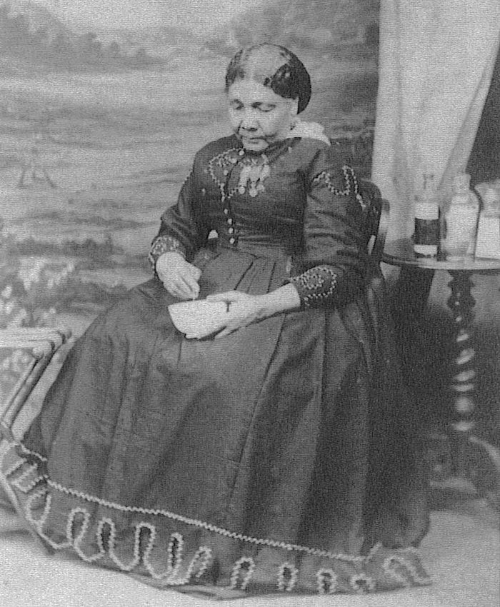 Black and white photograph of Mary Seacole, sitting tending to a small bowl, while wearing a fine dress with medals pinned to her chest
