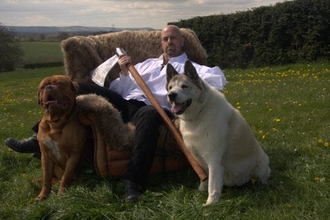 Author John Gwynne sitting on a fur-covered throne in a field, holding an axe with a large dog sitting on either side of him