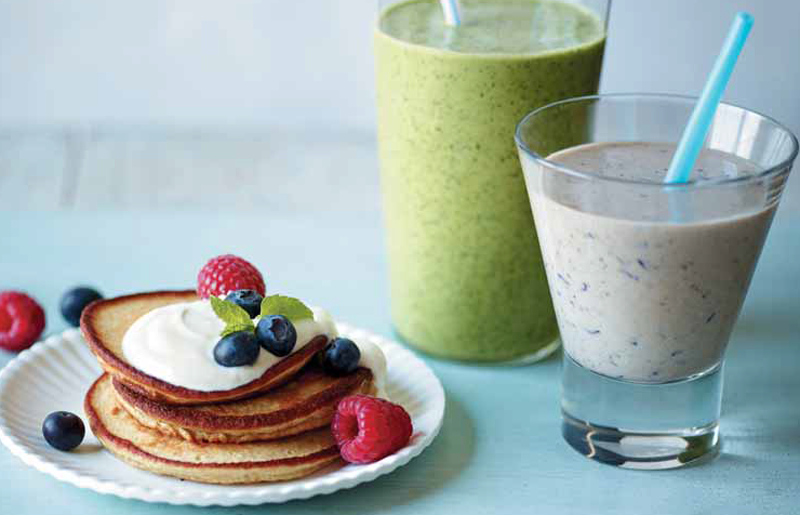 A pile of pancakes topped with Greek yoghurt, blueberries and raspberries next to two smoothies.