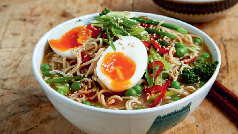 Close up photograph of ramen, showing noodles, brown broth, spring onions, chili peppers and a soft-boiled egg