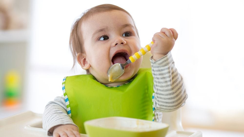 Baby sitting in a high chair with a green bib licking puree from a spoon