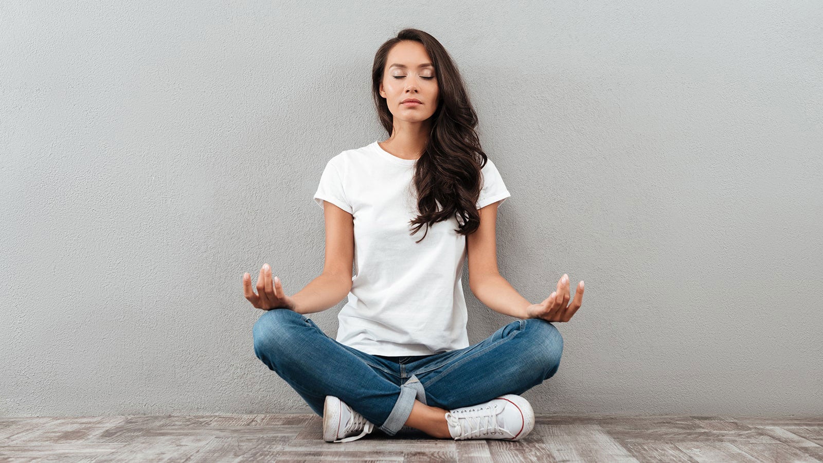 A woman dressed in white shirt and jeans sitting crossed legged, with her hands on her knees, in a classic yoga pose.