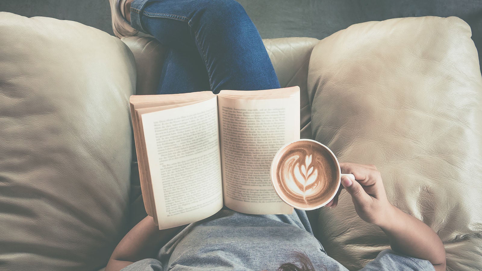 Image of a woman sitting on a sofa reading a book and holding a coffee