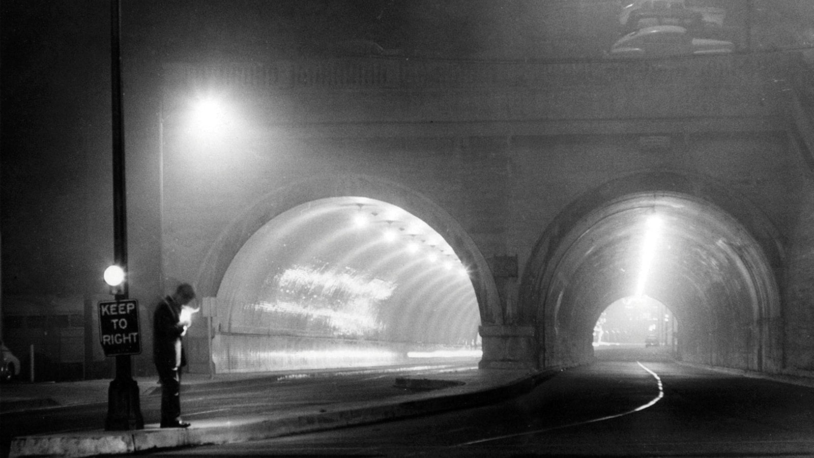 Black and white photo of man standing by the side of the road in front of two tunnels