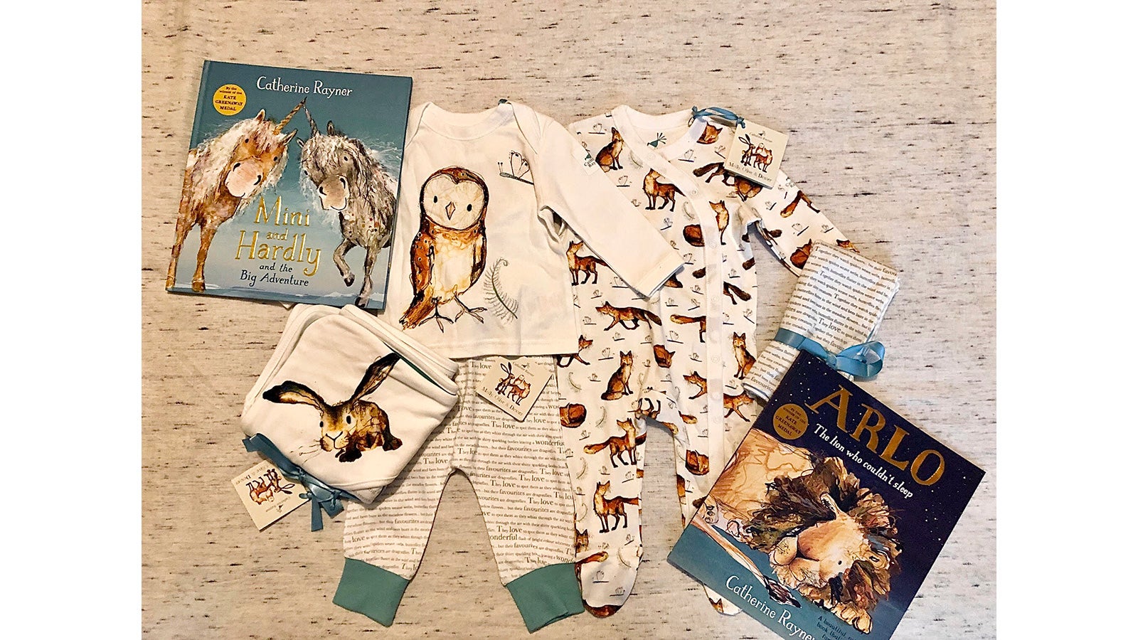 A bundle of babywear lies on a white patterned bedspread alongside a copy of Arlo the Lion Who Couldn't Sleep and Mini and Hardly and the Big Adventure