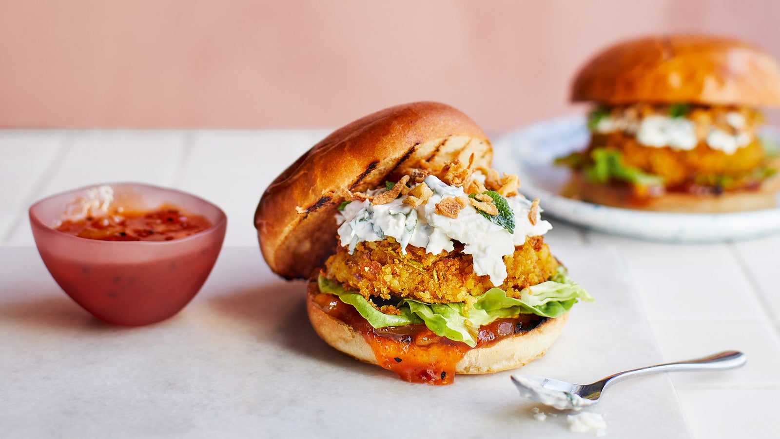 One chickpea burger served in a brioche bun with lettuce, relish and caulflower raita, nextto a small put of relish, with another chickpea burger out of focus in the background