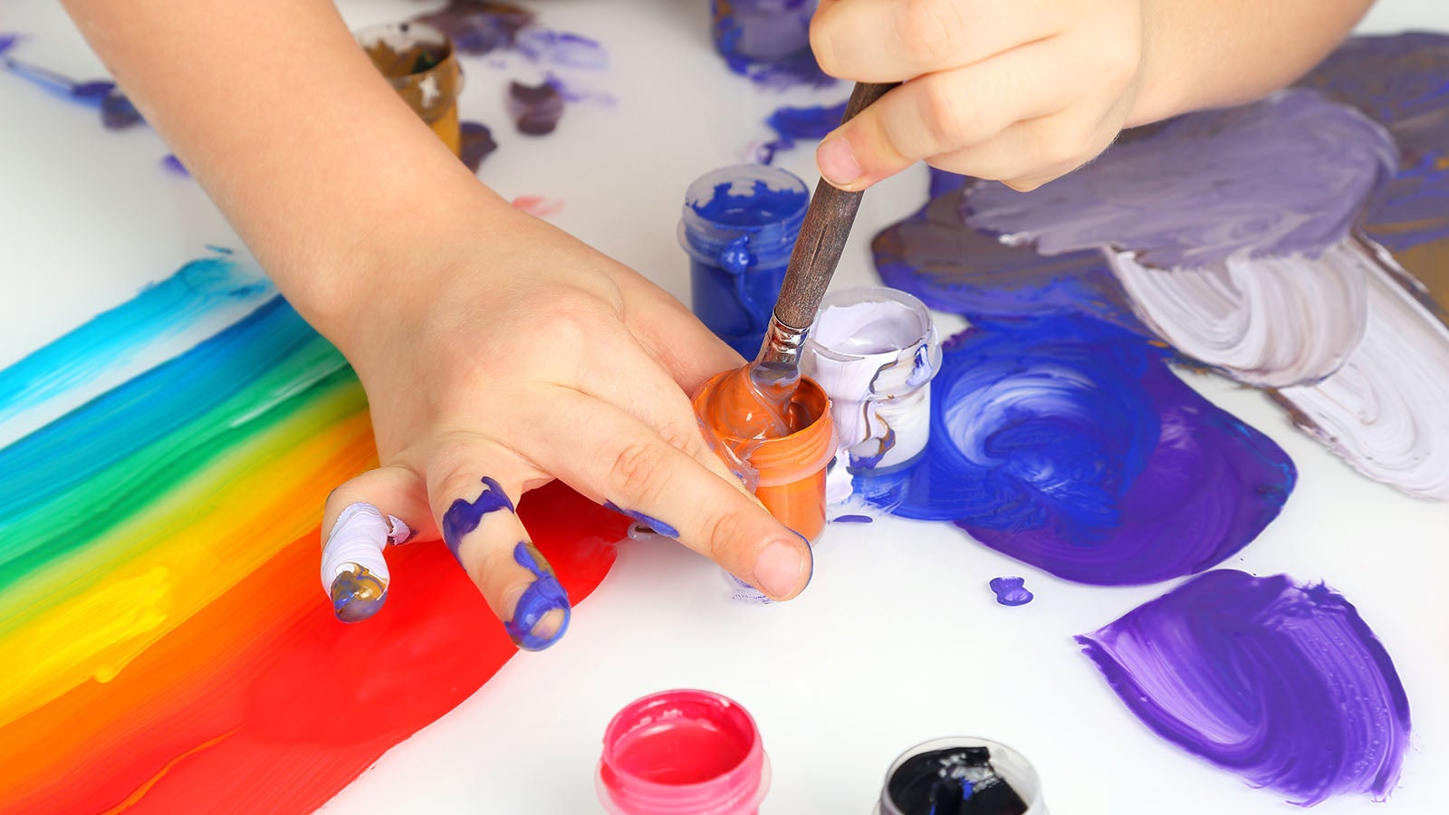 a close up of a child painting, a rainbow on paper beneath their hands, dipping a brush in a pot