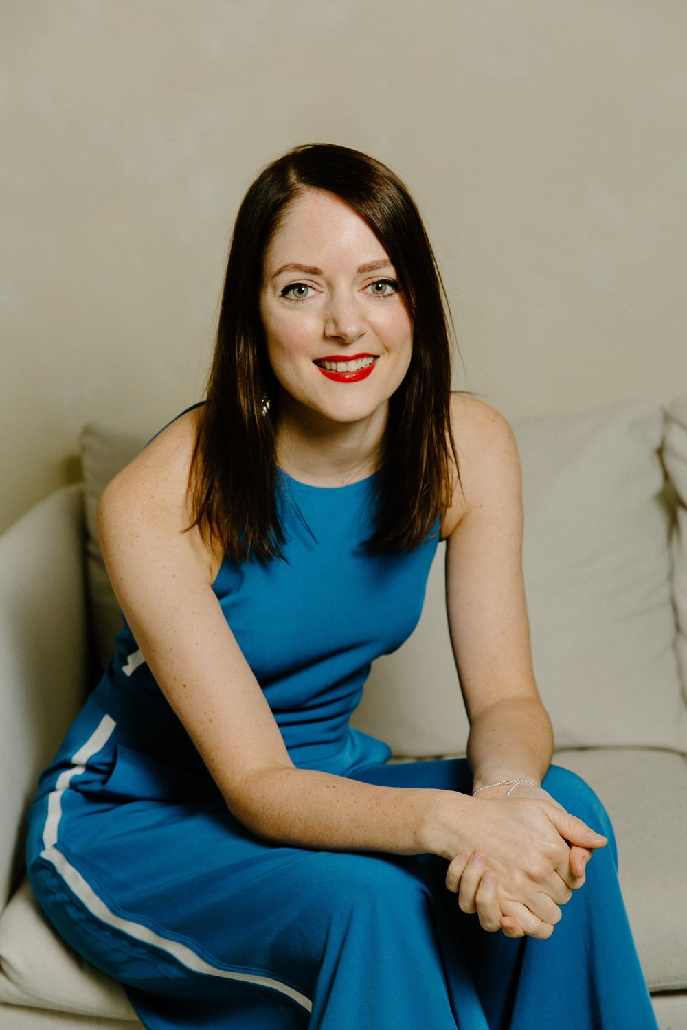 Laura Price wears a blue jumpsuit, sitting on a cream sofa and smiling to camera