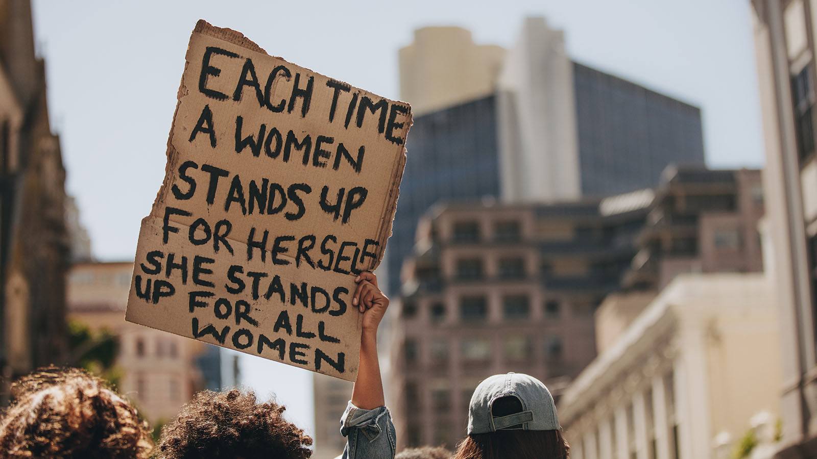 Image of a protest with a hand holding up a sign saying 'Each time a woman stands up for herself she stands up for all women.'