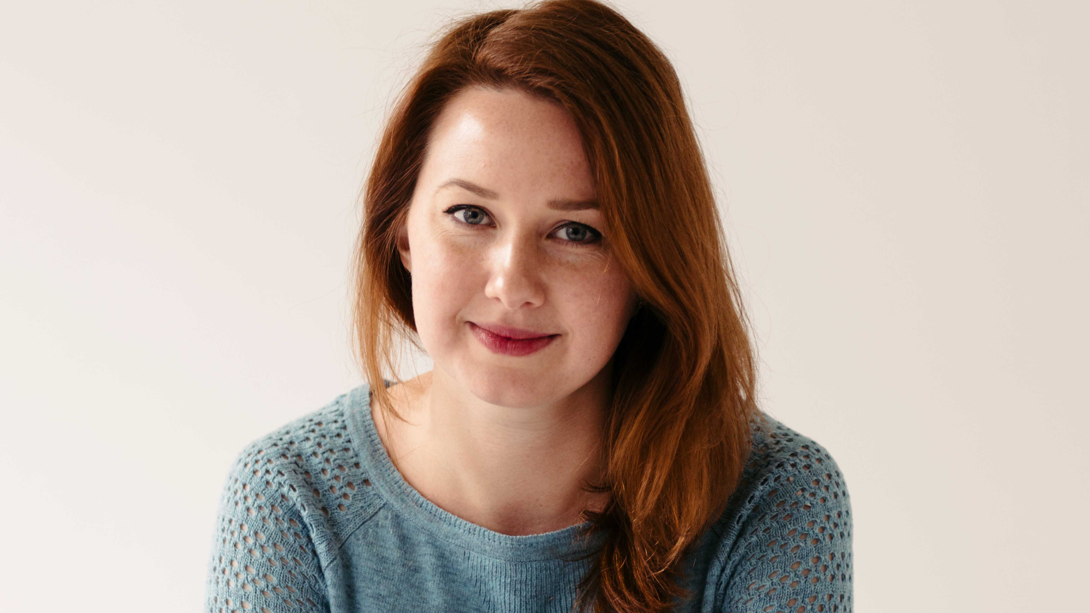 A photograph of Hannah Kent smiling at the camera wearing a blue jumper in front of a plain cream backdrop