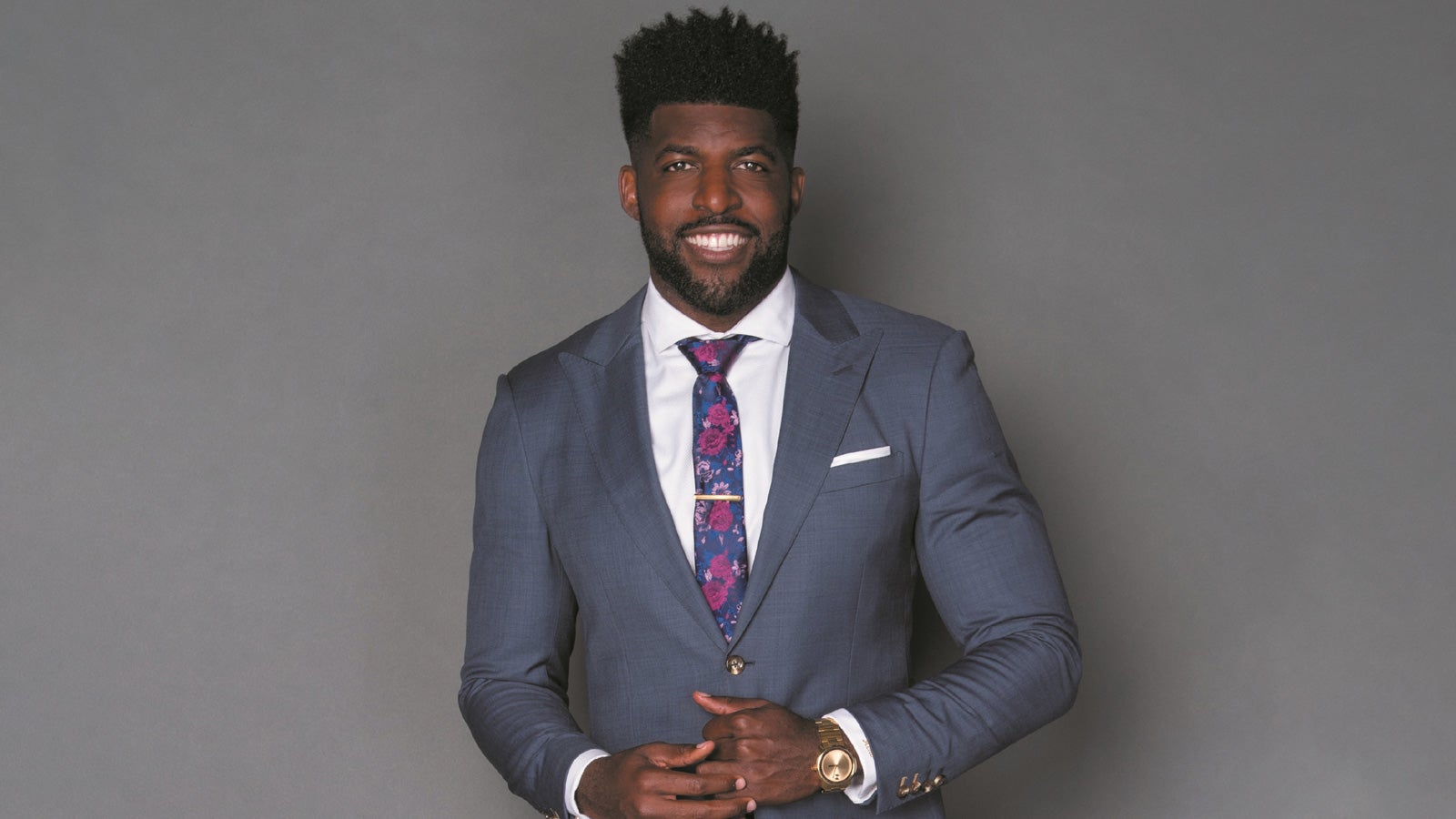 Emmanuel Acho smiling in a blue/grey suit in front of a grey background