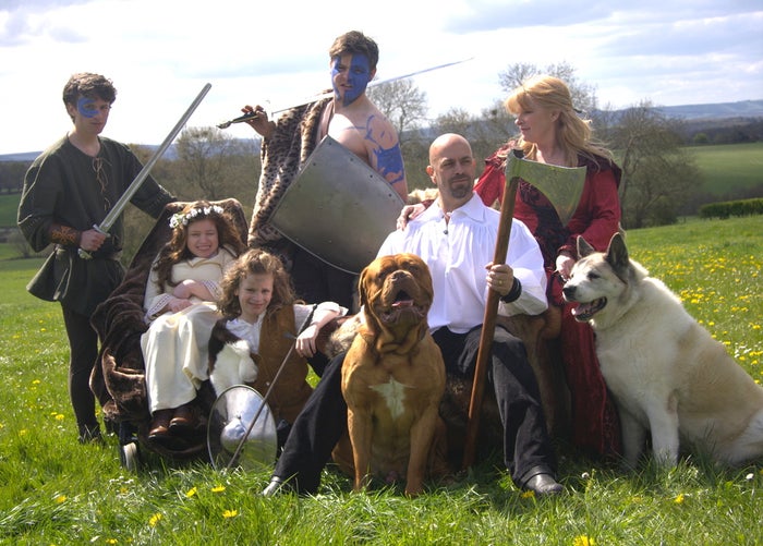 John Gwynne sitting on a fur-covered throne in a sunning field, holding an axe, surrounded by how two sons dresses as warriors, his wife, two daughters and two large dogs