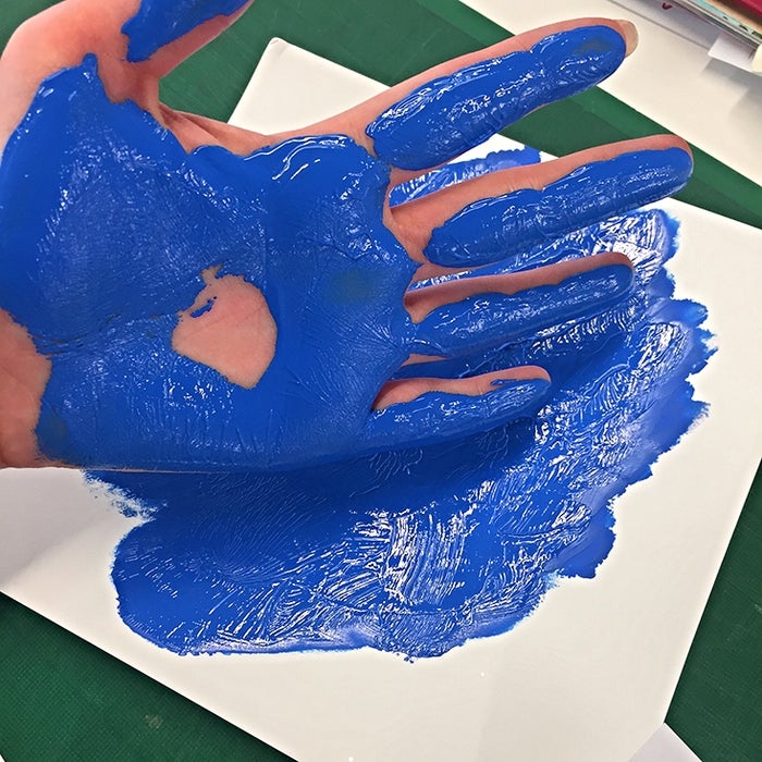 A hand covered in blue poster paint that has been used to leave a flower design on a piece of card