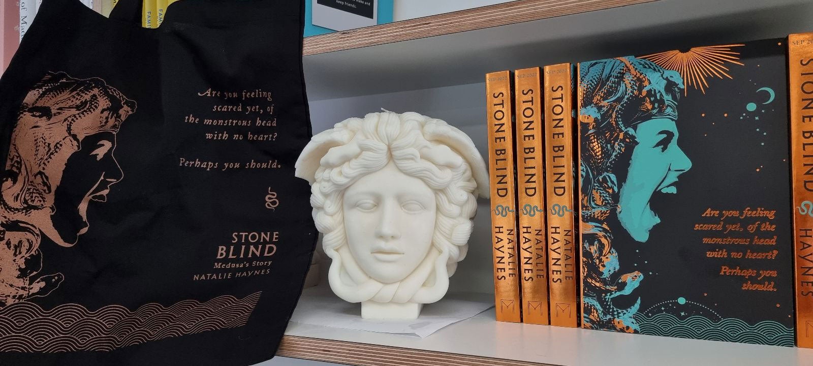 Proof copies of Stone Blind sit on a book shelf alongside a marble Medusa head and a Stone Blind themed tote bag.