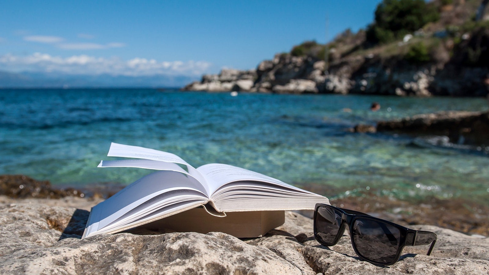 Book open next to sunglasses in front of blue sea