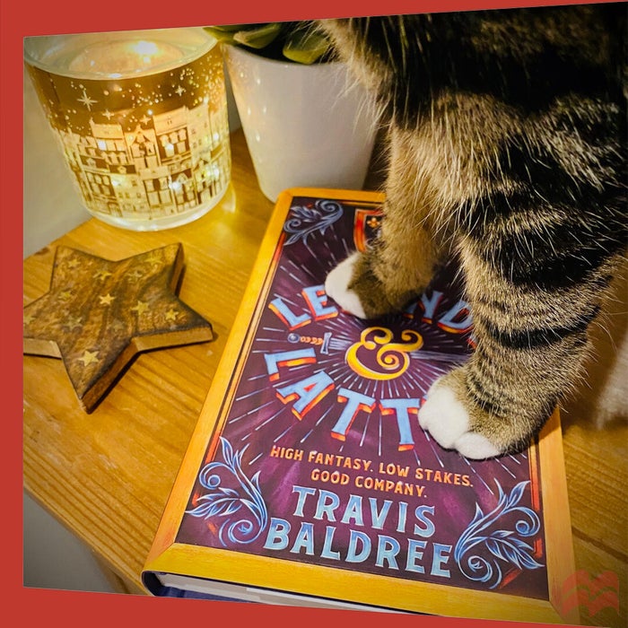 Two furry paws stand atop a copy of Legends & Lattes by Travis Baldree, preventing any reading from happening.