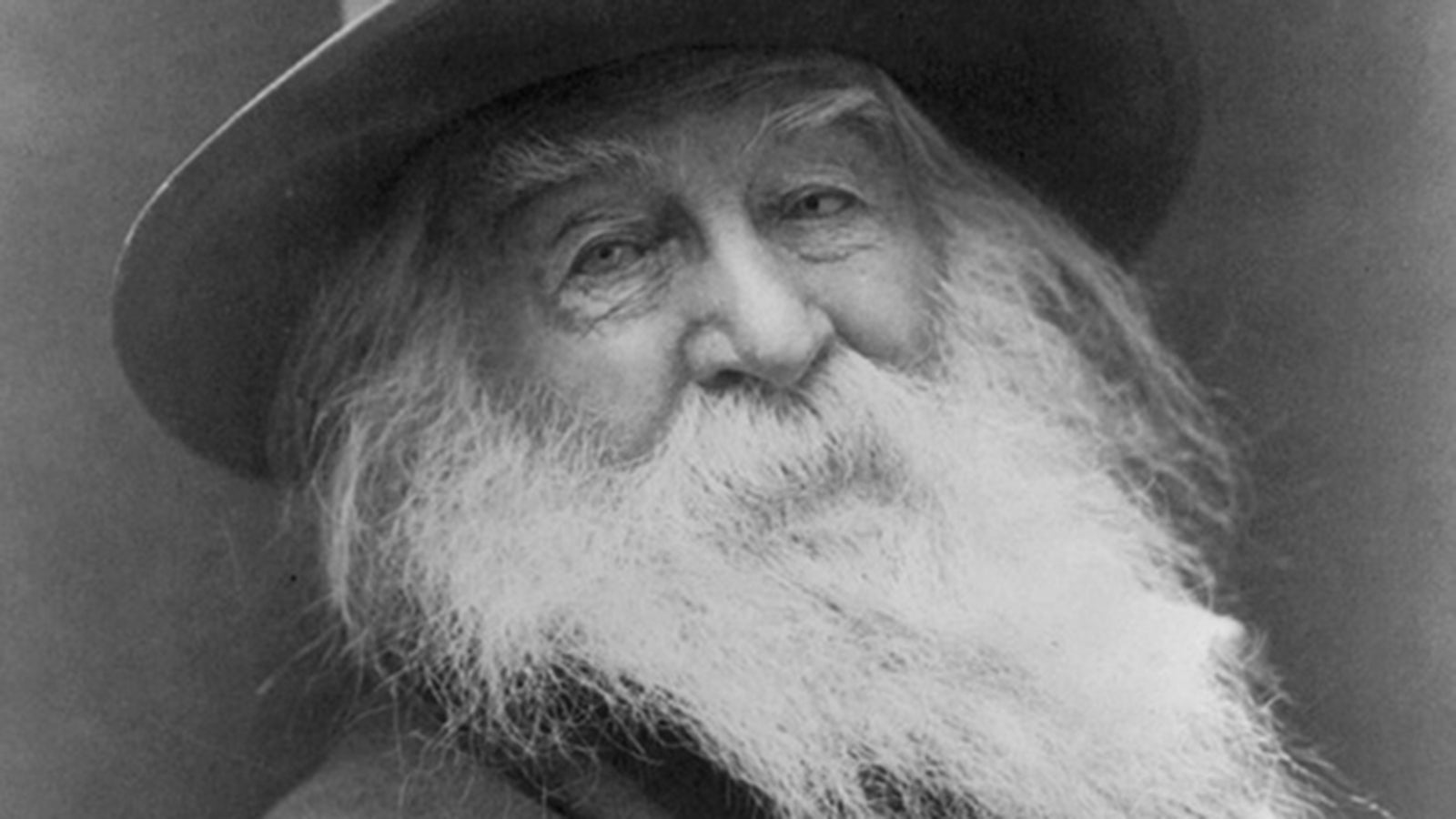 A close up black and white photograph of an elderly Walt Whitman, with a long white beard, wearing a dark wide-brimmed hat