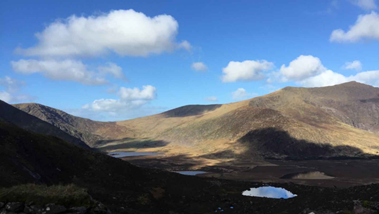 A landscape scene of mountains and a valley with small lakes within in, and clouds above in a bright blue sky casting shadows on the valley beneath.