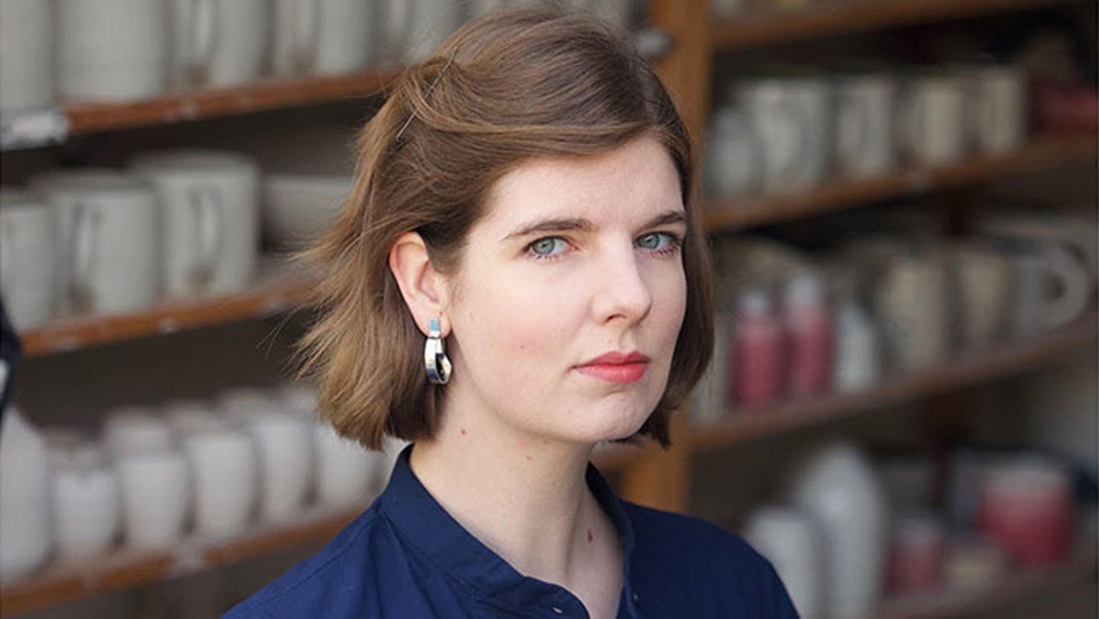 Elizabeth Macneal wears a blue shirt and looks to camera, standing in front of shelves full of ceramics