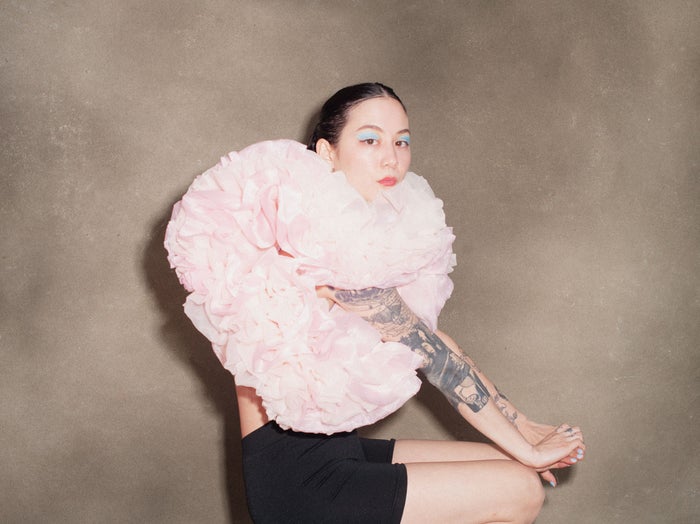 Michelle Zauner sitting sideways on a stool, wearing an oversized pink ruffled top, black shorts and bright blue eye shadow