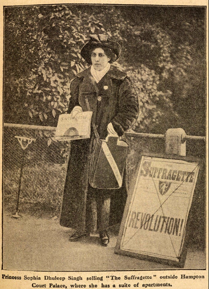 Sophia Duleep Singh standing next to a sign that says 'SUFFRAGETTE REVOLUTION' holding informational papers on the suffragette movement to hand out to the public