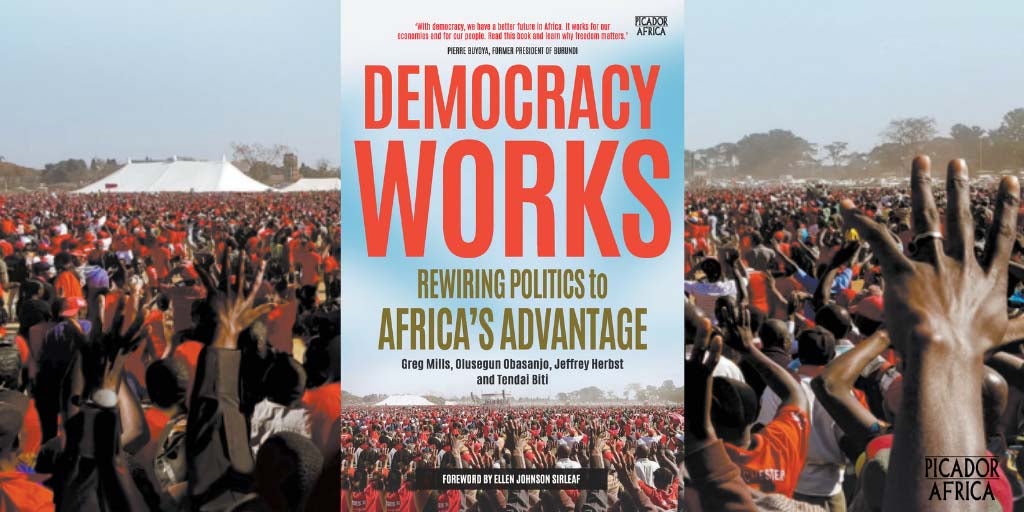 Photo of the book Democracy Works superimposed on a photo at a political gathering with hundreds of people with their hands in the air.
