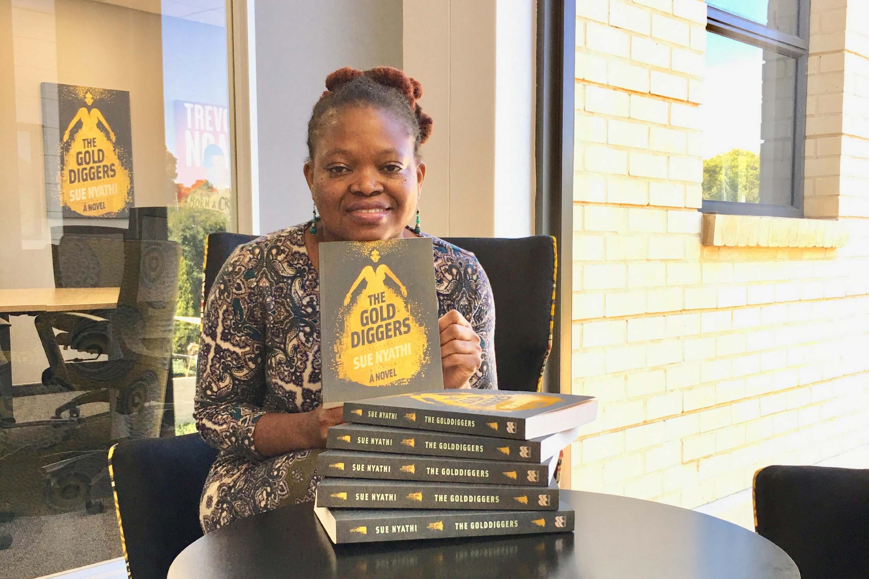 Photo of Sue Nyathi sat a table with copies of her book the The Gold diggers