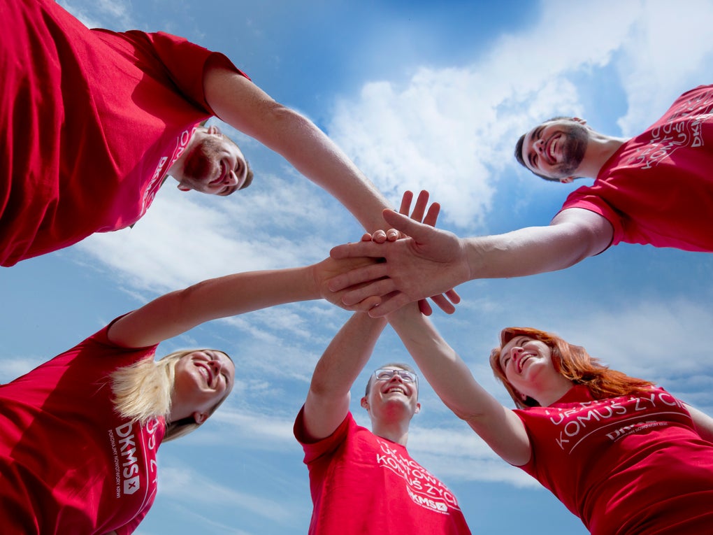 A group of five people in red DKMS shirts putting their hands in the middle together. You can see them from below. The sky above them is blue with some clouds and sunny weather. A group of five people in red DKMS shirts putting their hands in the middle together. You can see them from below. The sky above them is blue with some clouds and sunny weather.