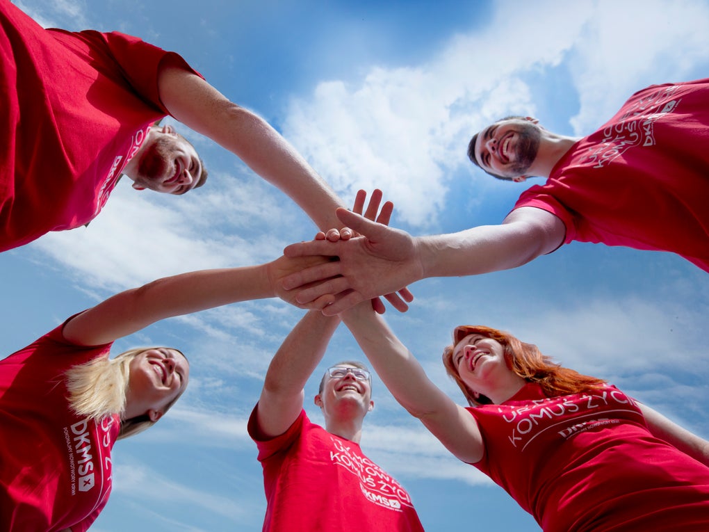 A group of five people in red DKMS shirts putting their hands in the middle together. You can see them from below. The sky above them is blue with some clouds and sunny weather. A group of five people in red DKMS shirts putting their hands in the middle together. You can see them from below. The sky above them is blue with some clouds and sunny weather.
