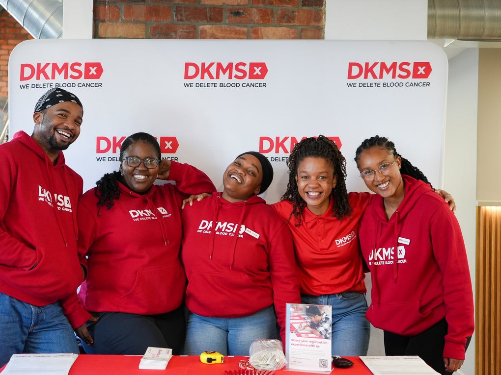 A group of five smiling DKMS team members stand together in front of a branded DKMS banner wall. They are wearing matching red DKMS hoodies and shirts. In front of them is an information table with flyers, swab kits, and a display encouraging registration for stem cell donation. A group of five smiling DKMS team members stand together in front of a branded DKMS banner wall. They are wearing matching red DKMS hoodies and shirts. In front of them is an information table with flyers, swab kits, and a display encouraging registration for stem cell donation.