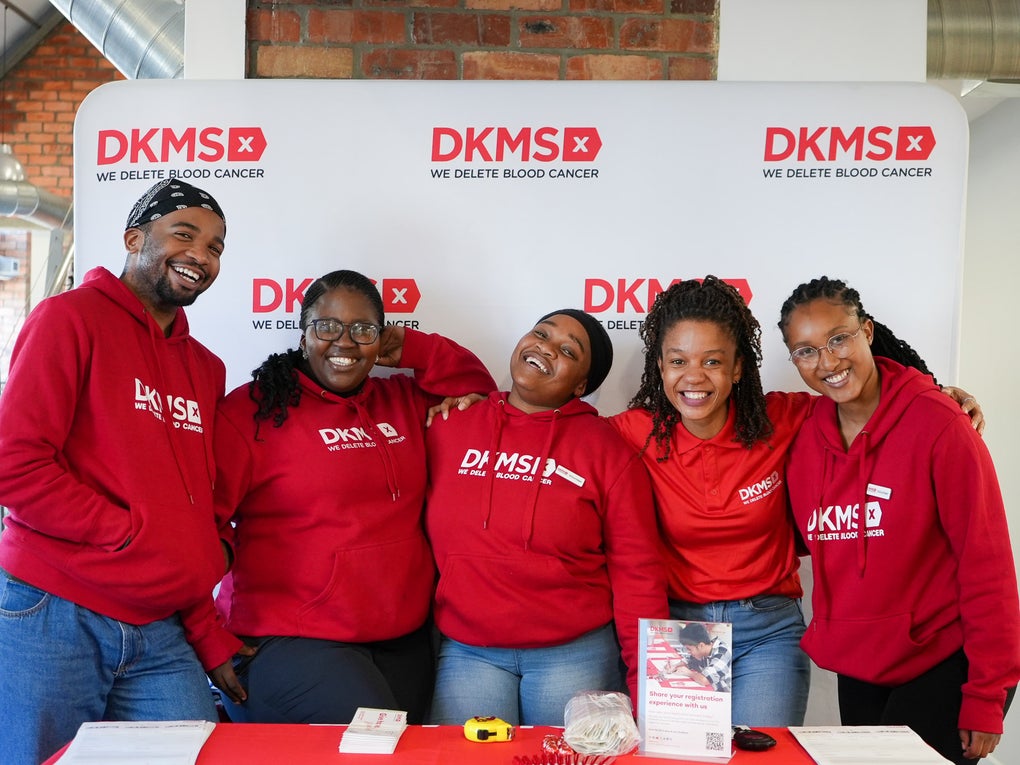 A group of five smiling DKMS team members stand together in front of a branded DKMS banner wall. They are wearing matching red DKMS hoodies and shirts. In front of them is an information table with flyers, swab kits, and a display encouraging registration for stem cell donation. A group of five smiling DKMS team members stand together in front of a branded DKMS banner wall. They are wearing matching red DKMS hoodies and shirts. In front of them is an information table with flyers, swab kits, and a display encouraging registration for stem cell donation.