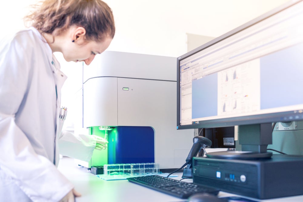 A young female scientist, wearing a white lab coat and gloves, is operating a flow cytometry machine in the DKMS Stem Cell Bank. A green light shines on the blood stem cell sample being analyzed, while a computer screen displays data and charts related to the quality of the blood stem cells. A young female scientist, wearing a white lab coat and gloves, is operating a flow cytometry machine in the DKMS Stem Cell Bank. A green light shines on the blood stem cell sample being analyzed, while a computer screen displays data and charts related to the quality of the blood stem cells.