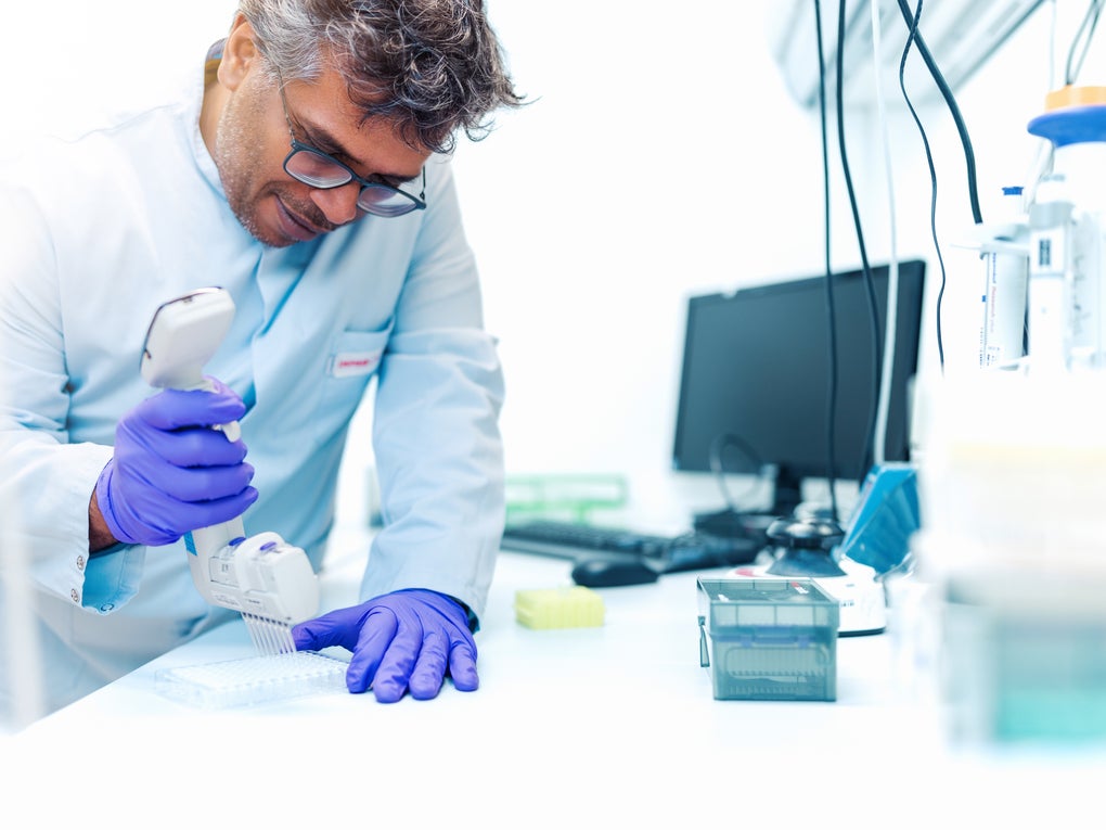 A male employee of the DKMS Life Science Lab, wearing a lab coat and purple gloves, uses a multichannel pipette to transfer samples into a well plate. Laboratory equipment and a computer are visible in the background. A male employee of the DKMS Life Science Lab, wearing a lab coat and purple gloves, uses a multichannel pipette to transfer samples into a well plate. Laboratory equipment and a computer are visible in the background.