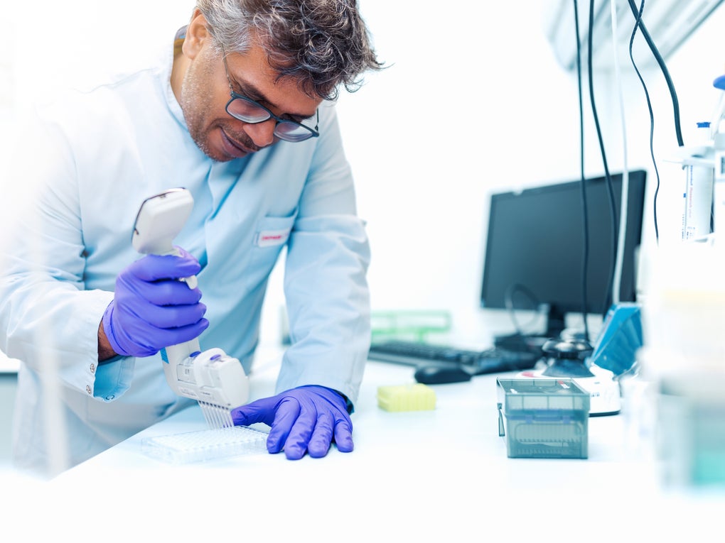 A male employee of the DKMS Life Science Lab, wearing a lab coat and purple gloves, uses a multichannel pipette to transfer samples into a well plate. Laboratory equipment and a computer are visible in the background. A male employee of the DKMS Life Science Lab, wearing a lab coat and purple gloves, uses a multichannel pipette to transfer samples into a well plate. Laboratory equipment and a computer are visible in the background.