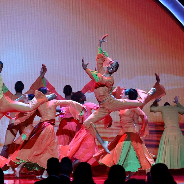 The Alvin Ailey Student Performance Group, wearing bright orange and pink costumes, perform an energetic dance performance on stage, with two dancers midair in dynamic jumps under colorful lighting. The Alvin Ailey Student Performance Group, wearing bright orange and pink costumes, perform an energetic dance performance on stage, with two dancers midair in dynamic jumps under colorful lighting.