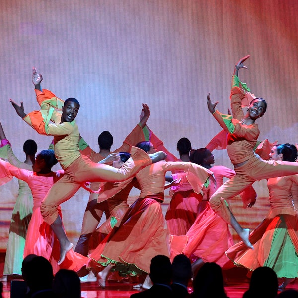 The Alvin Ailey Student Performance Group, wearing bright orange and pink costumes, perform an energetic dance performance on stage, with two dancers midair in dynamic jumps under colorful lighting. The Alvin Ailey Student Performance Group, wearing bright orange and pink costumes, perform an energetic dance performance on stage, with two dancers midair in dynamic jumps under colorful lighting.