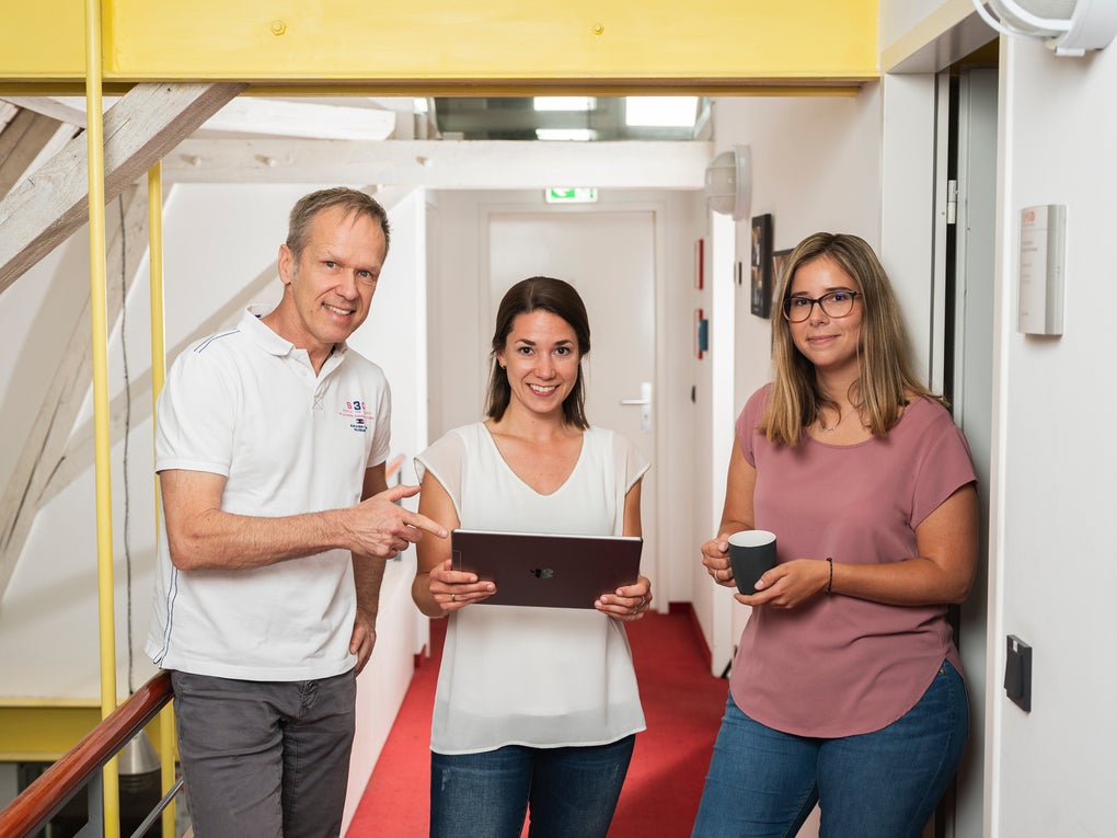 Three DKMS colleagues are standing together in a brigh hallway, smiling at the camera. The woman in the center is holding a laptop, while the man on the left points at the screen. The woman on the right is leaning against the wall, holding a coffee mug. Three DKMS colleagues are standing together in a brigh hallway, smiling at the camera. The woman in the center is holding a laptop, while the man on the left points at the screen. The woman on the right is leaning against the wall, holding a coffee mug.