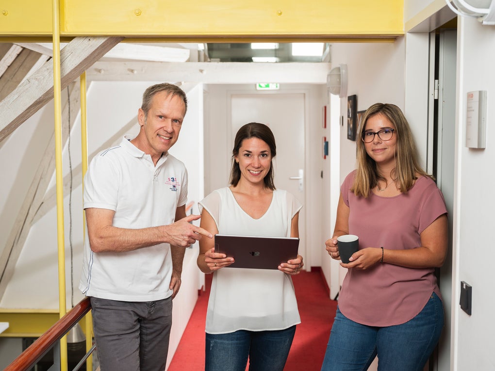 Three DKMS colleagues are standing together in a brigh hallway, smiling at the camera. The woman in the center is holding a laptop, while the man on the left points at the screen. The woman on the right is leaning against the wall, holding a coffee mug. Three DKMS colleagues are standing together in a brigh hallway, smiling at the camera. The woman in the center is holding a laptop, while the man on the left points at the screen. The woman on the right is leaning against the wall, holding a coffee mug.
