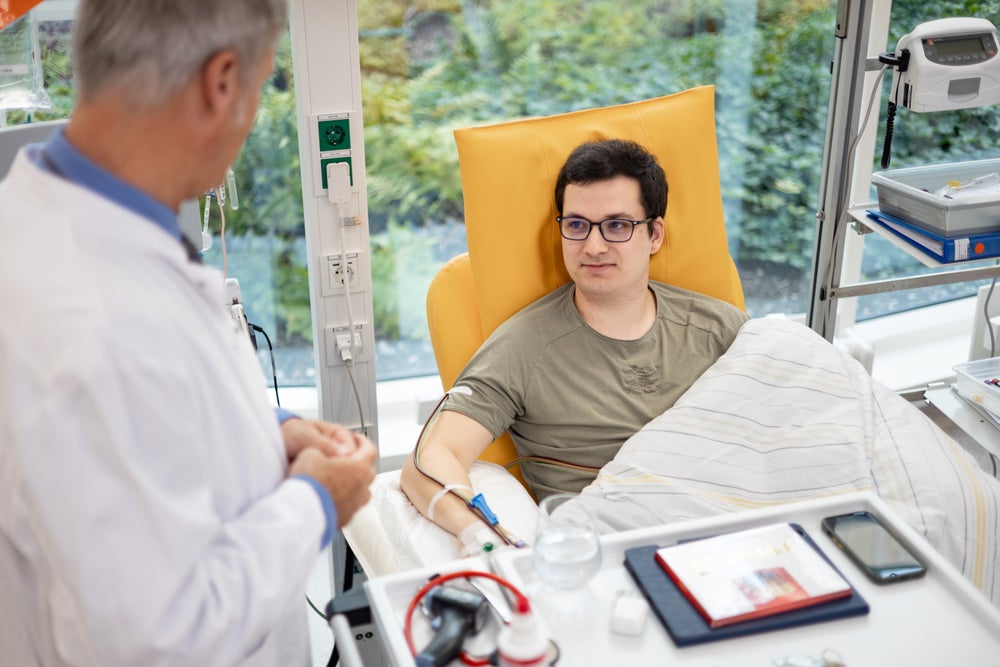 The young donor Lukas Hertzer is sitting in a yellow chair with a needle in his arm and connected to a apheresis tubing. He is the first donor whose cryopreserved stem cells were stored at the DKMS Stem Cell Bank. He is covered with a blanket and looking at Dr. Alexander Platz, the Medical Director of the Stem Cell Bank. In the background medical equipment and a big window with a view of the greenery outside are visible. The young donor Lukas Hertzer is sitting in a yellow chair with a needle in his arm and connected to a apheresis tubing. He is the first donor whose cryopreserved stem cells were stored at the DKMS Stem Cell Bank. He is covered with a blanket and looking at Dr. Alexander Platz, the Medical Director of the Stem Cell Bank. In the background medical equipment and a big window with a view of the greenery outside are visible.
