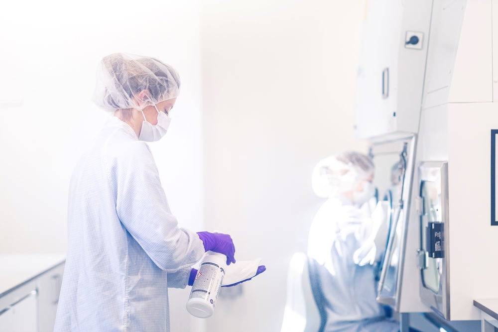 Two female laboratory technicians in protective clothing, including white lab coats, face masks, hairnets, and purple gloves, are working in a clean and sterile room of the DKMS Life Science Lab. One has a disinfectant spray and wipe in her hands, while the other is seated at a bioisolator, handling the aseptic production of a Adult Donor Cryopreserved Units (ADCU). Two female laboratory technicians in protective clothing, including white lab coats, face masks, hairnets, and purple gloves, are working in a clean and sterile room of the DKMS Life Science Lab. One has a disinfectant spray and wipe in her hands, while the other is seated at a bioisolator, handling the aseptic production of a Adult Donor Cryopreserved Units (ADCU).