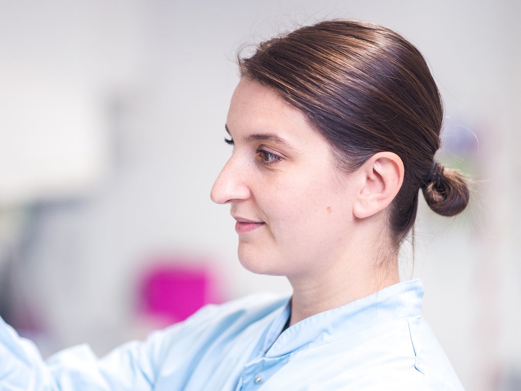A female employee of the Life Science Lab, with brown hair in a bun, wearing a light blue lab coat, and green protective gloves, is working in the laboratory. She is holding a yellow stack of sample containers and placing them into a machine. The background is blurred A female employee of the Life Science Lab, with brown hair in a bun, wearing a light blue lab coat, and green protective gloves, is working in the laboratory. She is holding a yellow stack of sample containers and placing them into a machine. The background is blurred