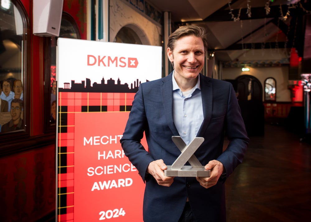 The winner of the Mechtild Harf Science Award Prof. Robert Zeiser, wearing a dark suit, holds the award trophy, in the shape of an x, and stands in front of a red DKMS banner that reads "Mechtild Harf Science Award 2024". The winner of the Mechtild Harf Science Award Prof. Robert Zeiser, wearing a dark suit, holds the award trophy, in the shape of an x, and stands in front of a red DKMS banner that reads "Mechtild Harf Science Award 2024".