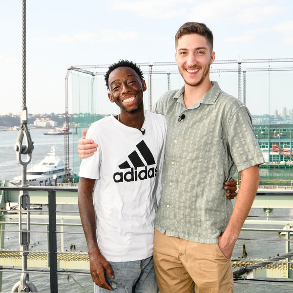 Two smiling young men, who are donor and patient, stand side by side on a platform with their arms around each other. The guy on the left wears a white Adidas t-shirt, the other one on the right a light green patterned shirt and beige shorts. In the background, a river, boats, and a city skyline are visible. Two smiling young men, who are donor and patient, stand side by side on a platform with their arms around each other. The guy on the left wears a white Adidas t-shirt, the other one on the right a light green patterned shirt and beige shorts. In the background, a river, boats, and a city skyline are visible.